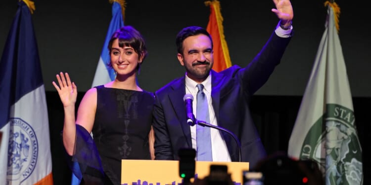 A man and woman standing on a stage with a microphone and a flag behind them