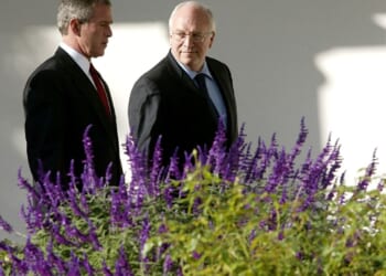 A pair of men in suits standing next to purple flowers