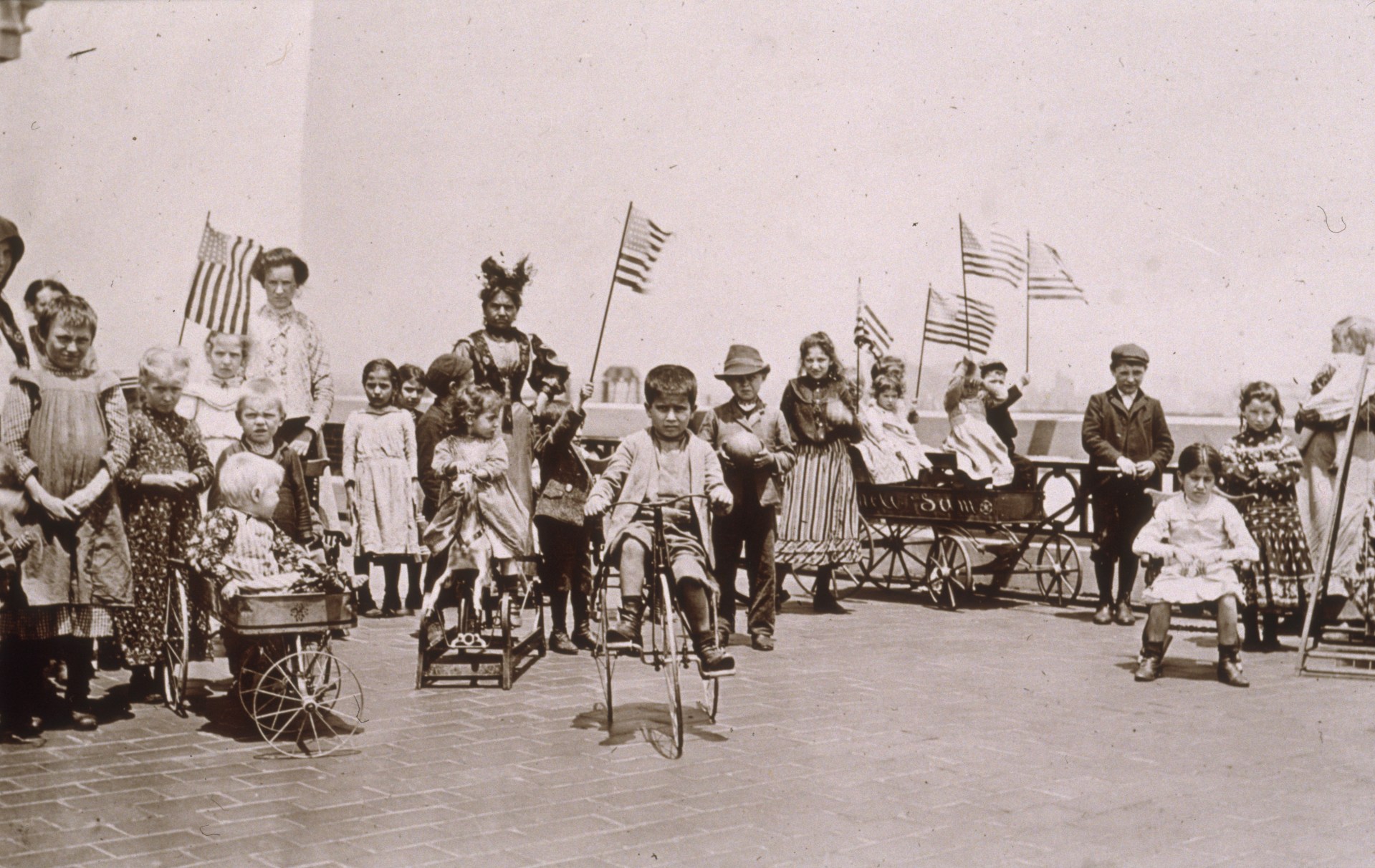Children holding American flags while riding tricycles and wagons on the rooftop garden of Ellis Island, New York City. (Photo by Jacob A. Riis/Museum of the City of New York/Getty Images)