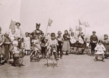 Children holding American flags while riding tricycles and wagons on the rooftop garden of Ellis Island, New York City. (Photo by Jacob A. Riis/Museum of the City of New York/Getty Images)