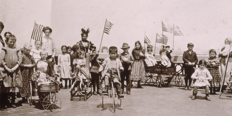 Children holding American flags while riding tricycles and wagons on the rooftop garden of Ellis Island, New York City. (Photo by Jacob A. Riis/Museum of the City of New York/Getty Images)