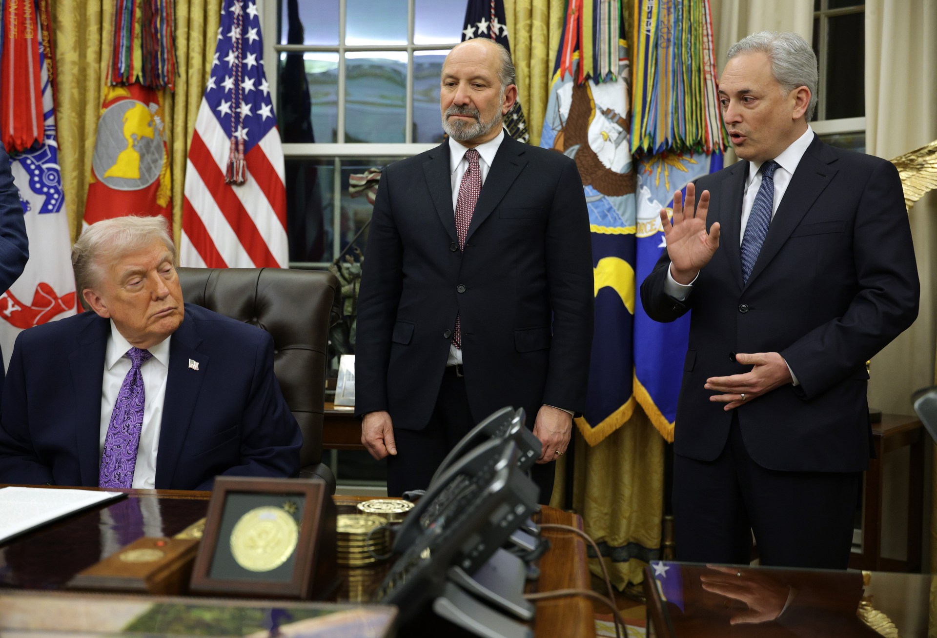A man in a dark suit with a purple tie sits at a desk in an ornate office with military flags and the American flag in the background, while two men in business suits stand nearby, one gesturing while speaking.