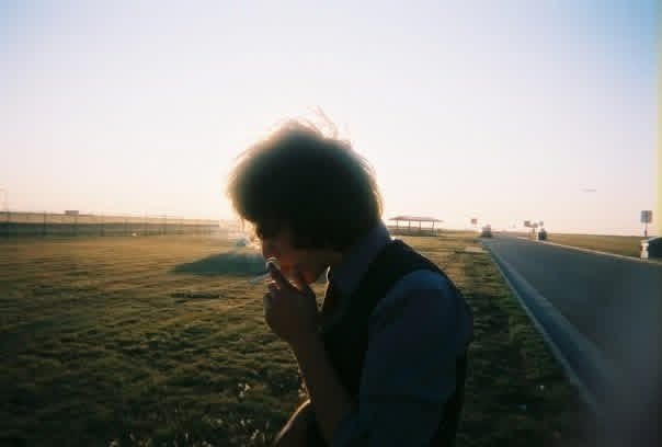 A person with curly hair, wearing a dark jacket, stands in profile on a flat field during golden hour, with their hand near their face in a contemplative pose.