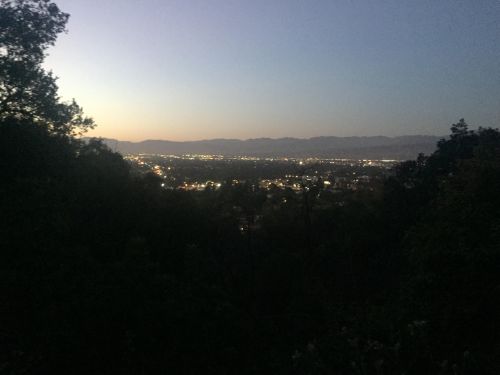 A valley town nestled between forested hills at dusk, with scattered lights illuminating homes across the landscape as the sky transitions from daylight to evening.