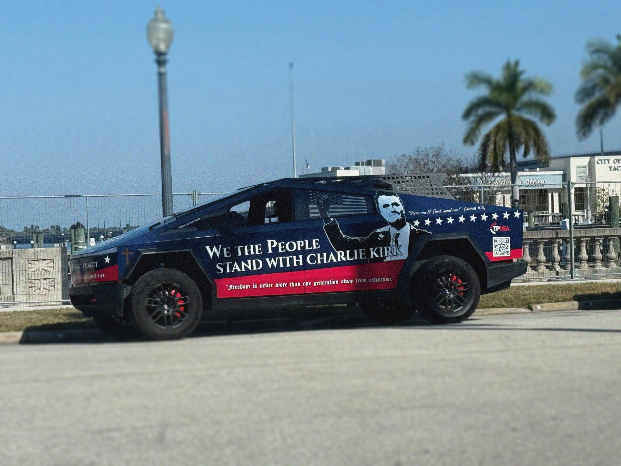 A dark blue pickup truck wrapped with political messaging and imagery is parked in a waterfront area with palm trees and buildings visible in the background.