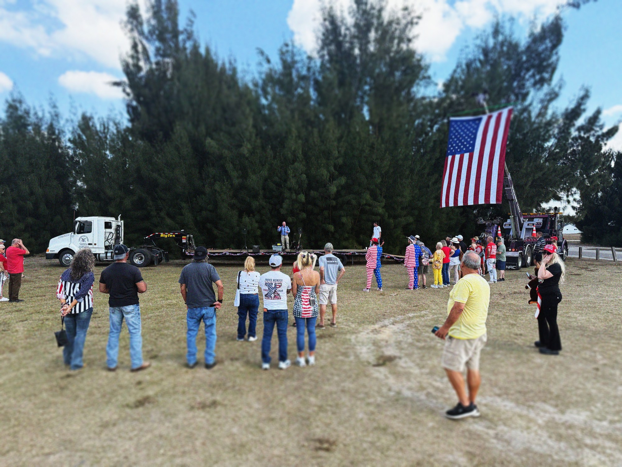 A crowd gathers in an open field with tall evergreen trees in the background to watch a large American flag displayed on a truck during a patriotic outdoor event.