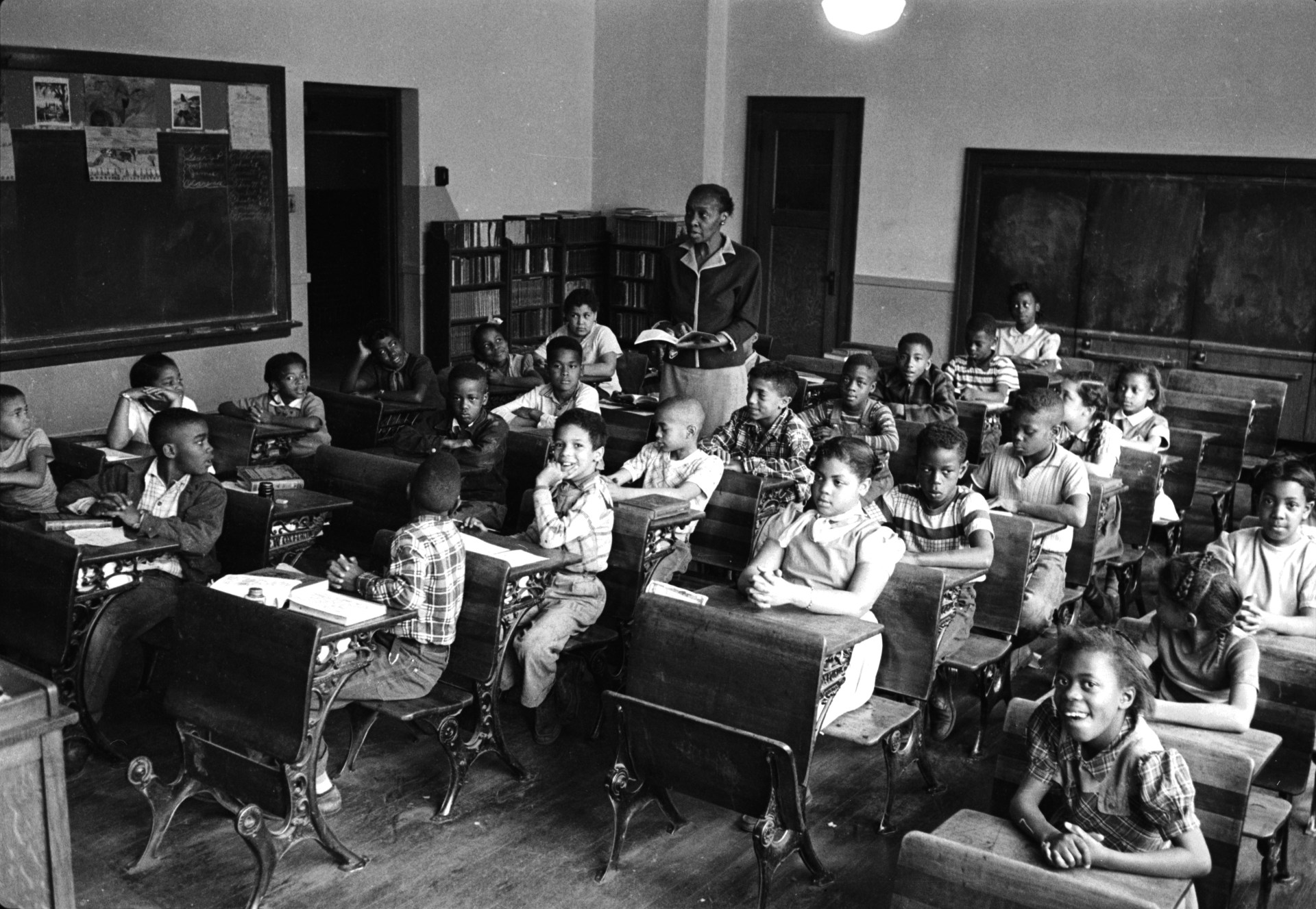 A teacher instructs a diverse classroom of young students seated at desks in a mid-20th century schoolroom with blackboards and bookshelves.