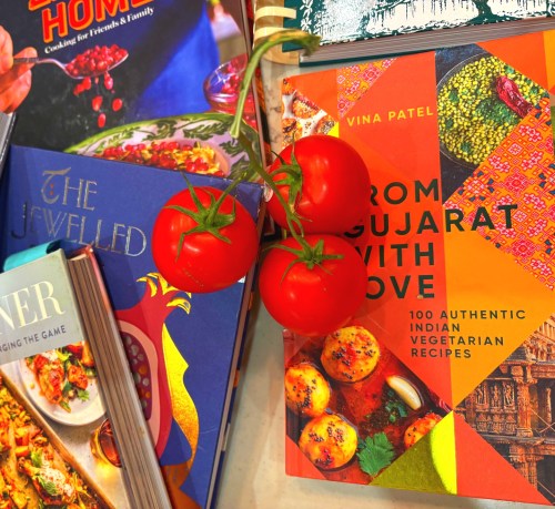 Fresh tomatoes on the vine rest atop a collection of Indian vegetarian cookbooks displayed on a shelf, including titles by Vina Patel and others featuring colorful food photography and cultural patterns.