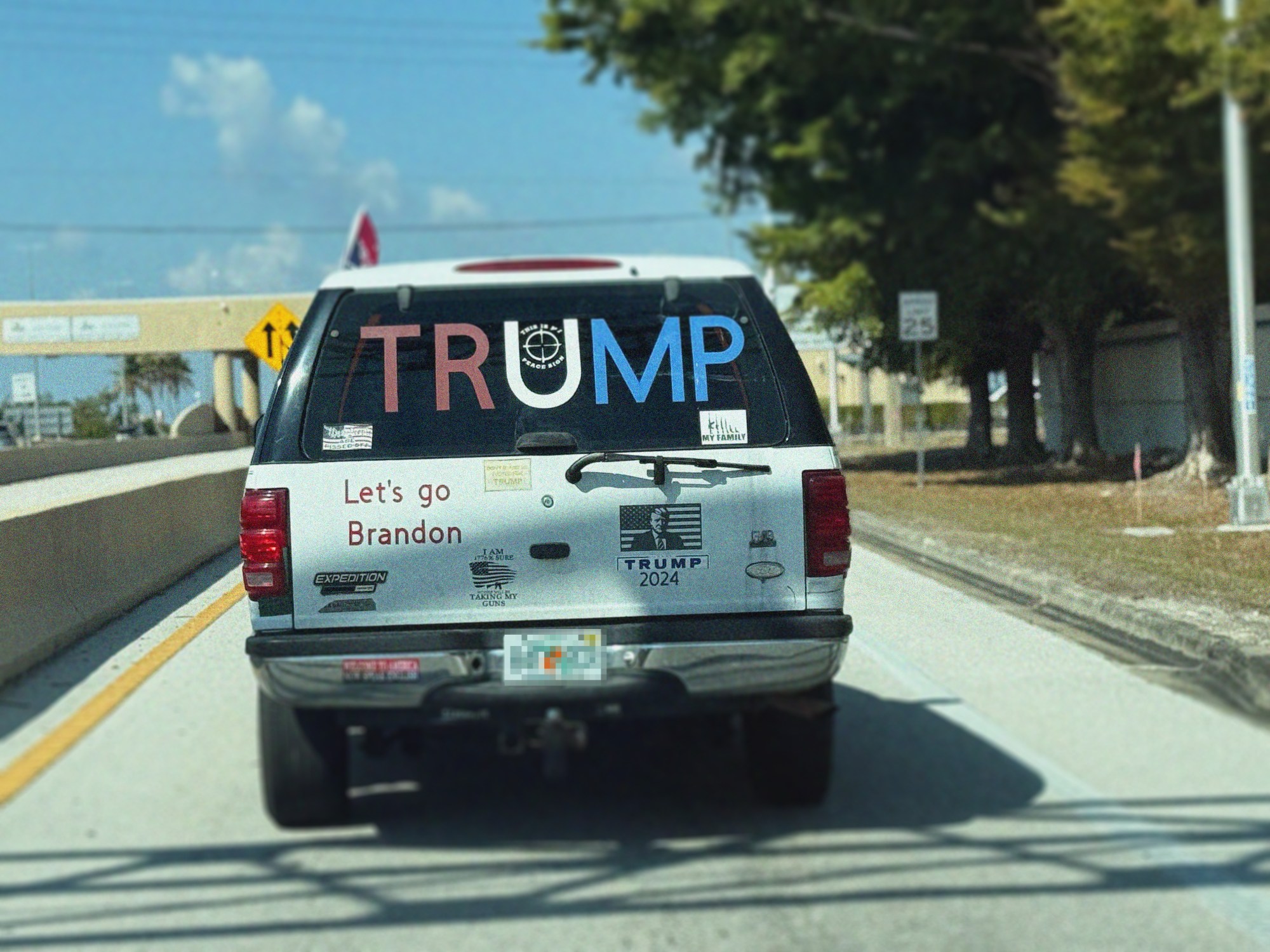 A white and black Ford Expedition with large "TRUMP" lettering and political slogans including "Let's go Brandon" and "TRUMP 2024" is driven on a highway during daytime.