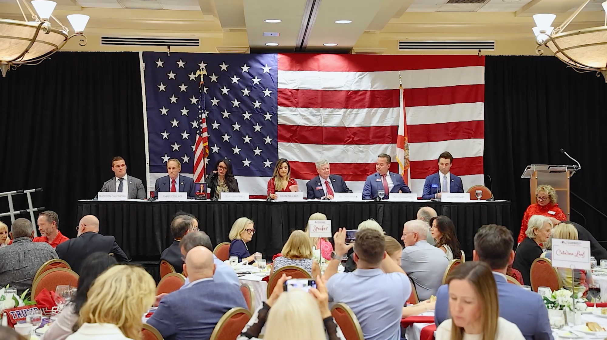 Seven panelists sit at a table on stage behind American flags during a formal event, addressing an audience seated at round tables in an elegant ballroom setting.