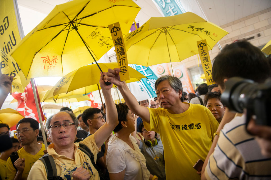 A crowd of protesters wearing yellow clothing and holding yellow umbrellas gather indoors, with Chinese characters visible on their shirts and umbrellas.