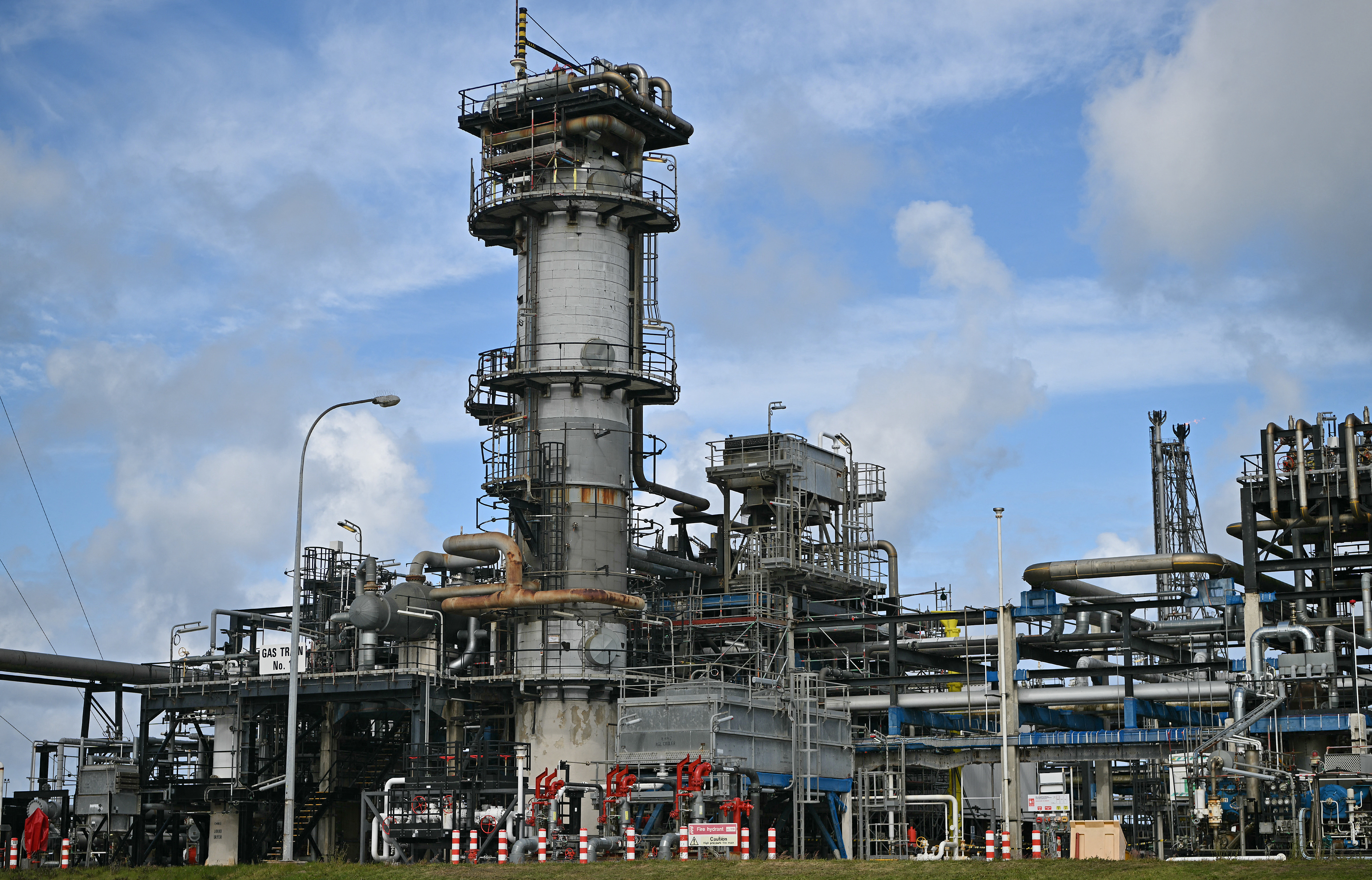 An industrial refinery complex with a tall distillation tower, interconnected piping systems, and processing equipment under a partly cloudy sky.