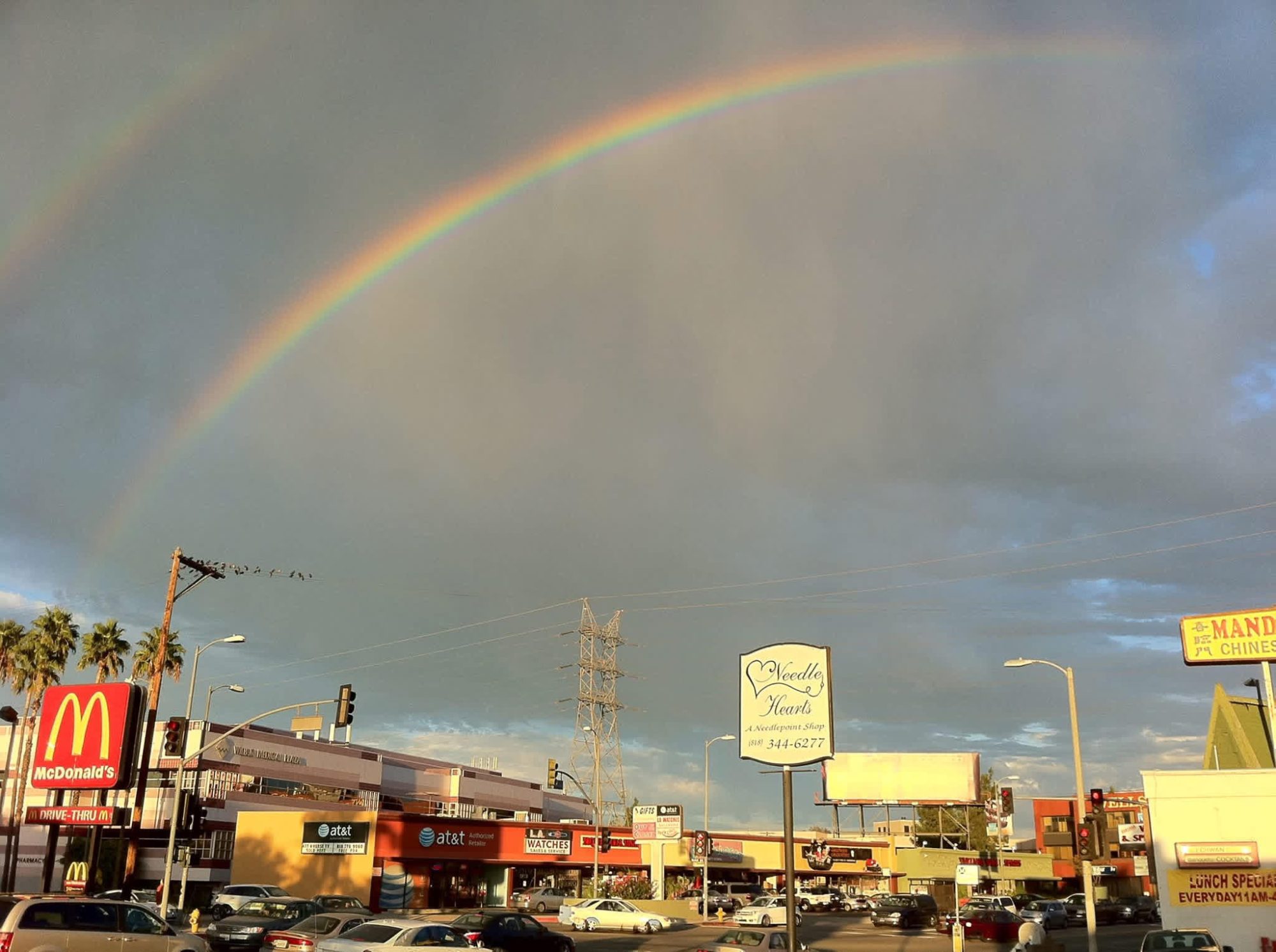 A vibrant rainbow arcs over a commercial strip mall with McDonald's, AT&T, and other businesses visible along a busy street lined with palm trees and power lines.