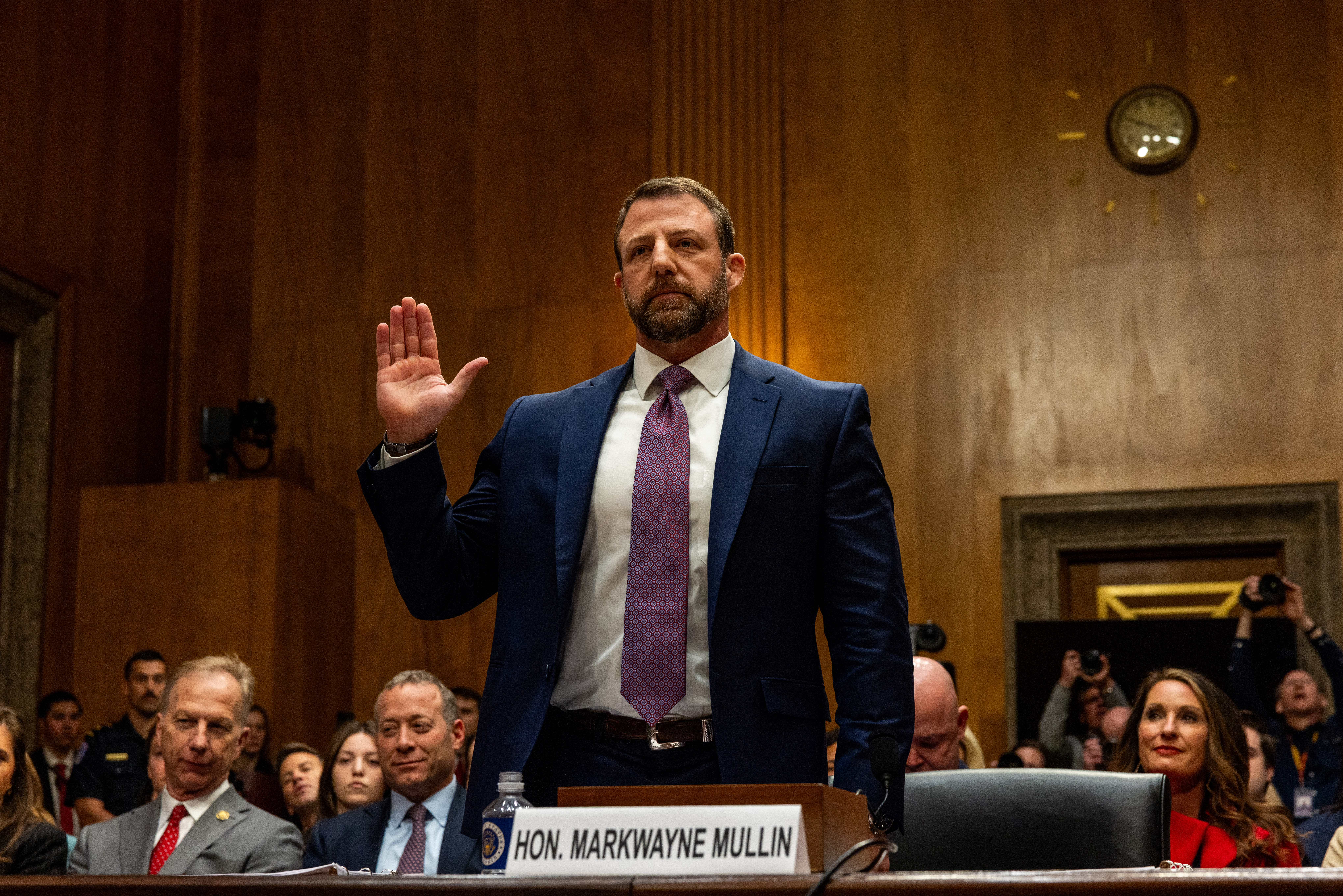A man in a blue suit and purple tie raises his hand while standing at a witness table during what appears to be a formal congressional hearing in a wood-paneled chamber.