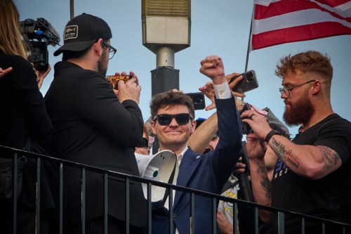 A man in sunglasses and a blue suit raises his fist while surrounded by people recording on phones, with an American flag visible in the background at what appears to be a public outdoor event.