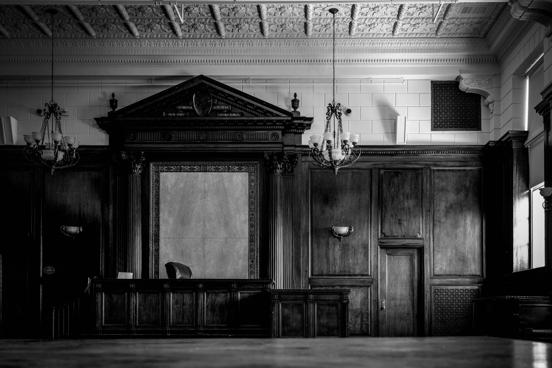 A grand neoclassical courtroom with ornate black wood paneling, classical columns, decorative pediment bearing Latin inscriptions, hanging chandeliers, and a judge's bench, photographed in black and white.