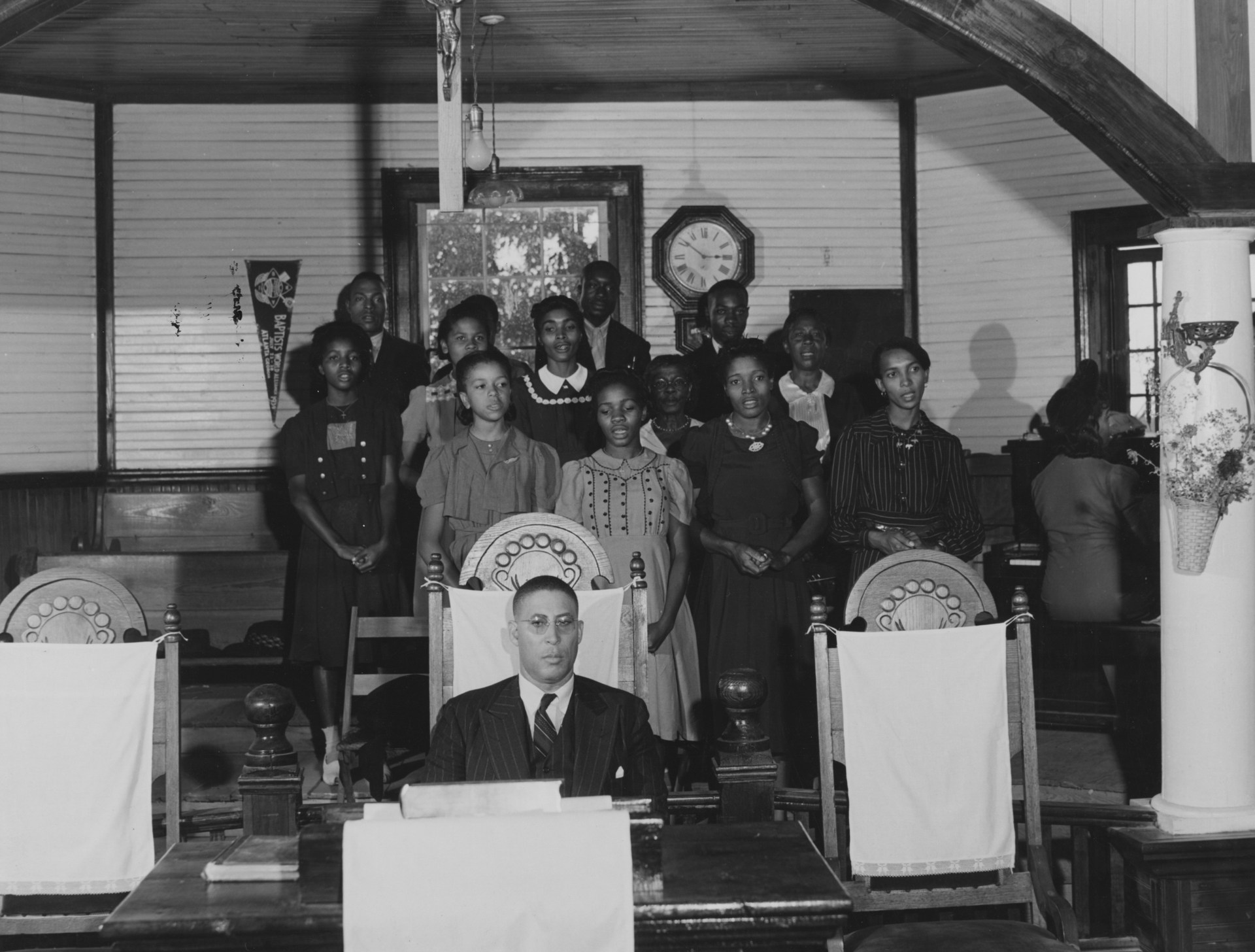 A Black man in a suit sits at a desk in a formal interior space while a group of Black visitors stands behind him in what appears to be an official or institutional setting from the mid-20th century.