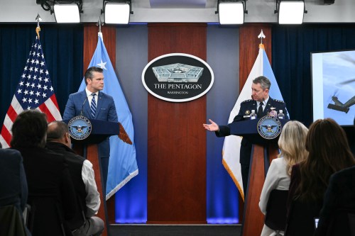 A military officer and civilian official speak at podiums during a press conference in The Pentagon, with the U.S. flag and two other flags displayed behind them.