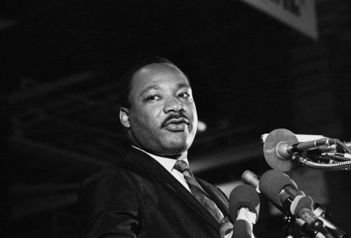 A man in a dark suit and tie speaks at multiple microphones during what appears to be a formal address or press conference, captured in black and white photography.
