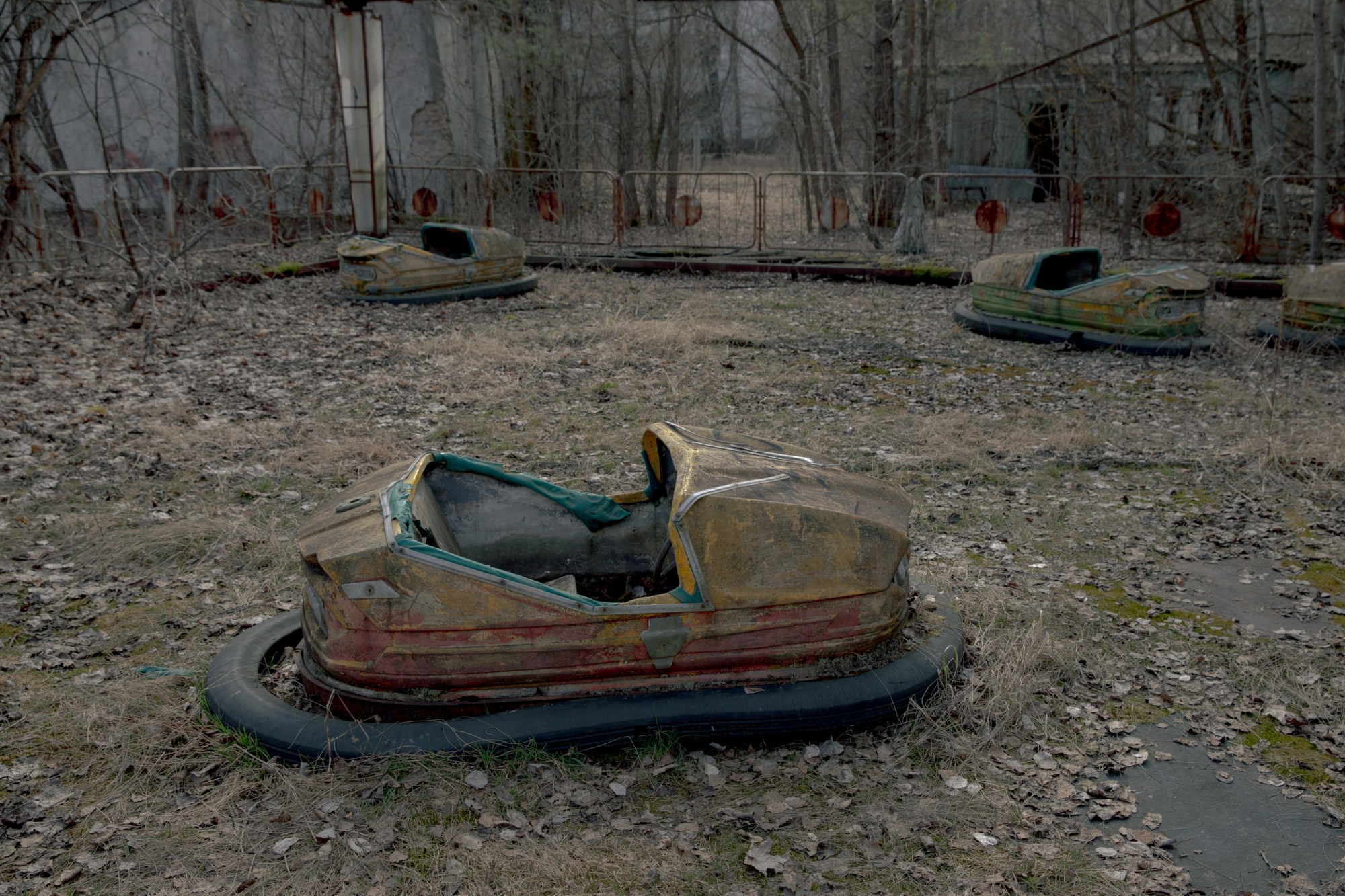 Abandoned, decaying bumper cars sit rusting on the ground of a derelict amusement park surrounded by bare trees and deteriorating fencing.