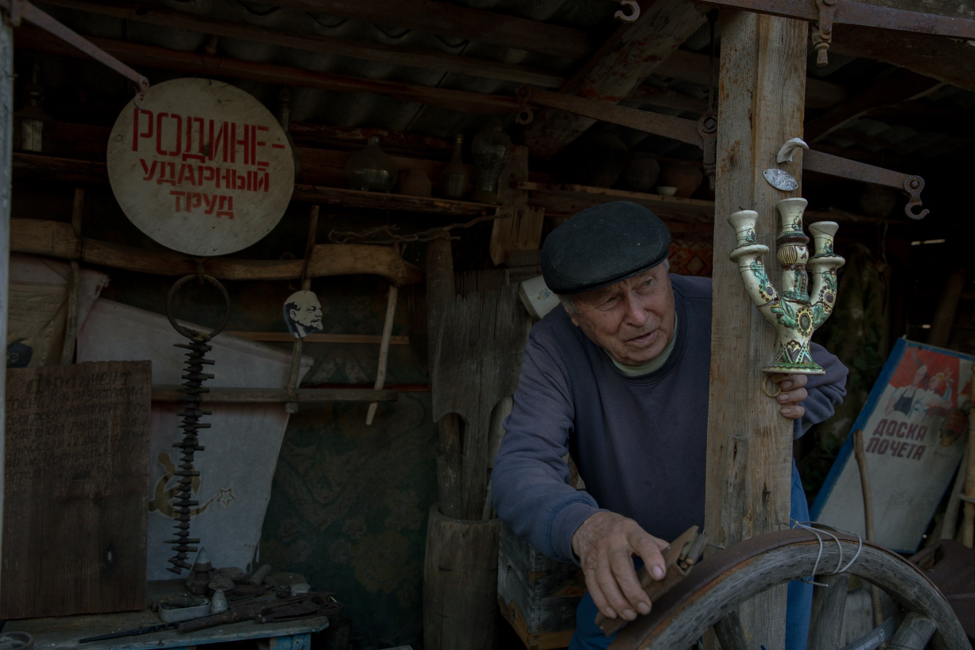 An elderly man in a dark cap works in a cluttered workshop filled with Soviet-era memorabilia, holding a decorative candelabra while standing among wooden beams and vintage items.