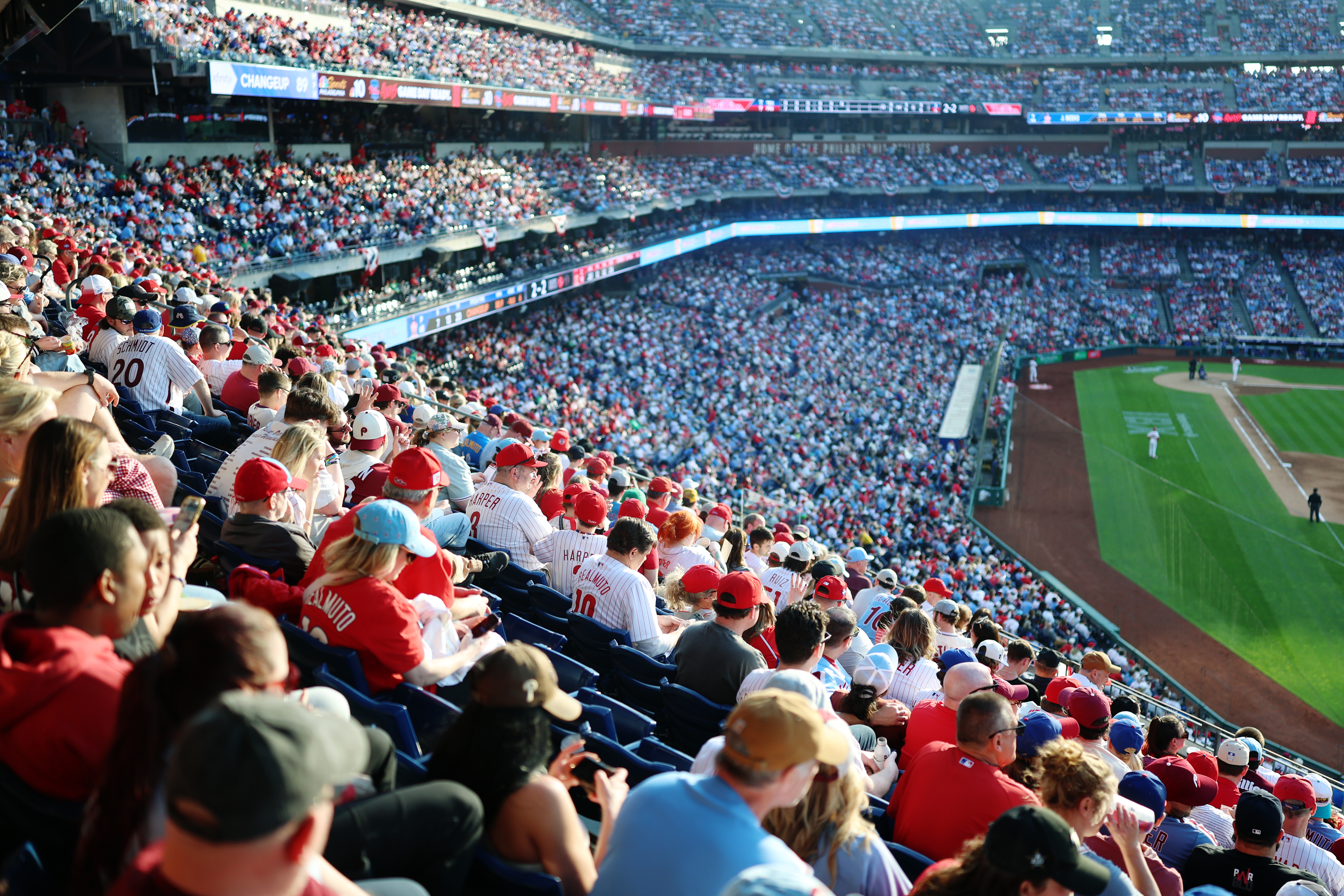 A packed baseball stadium filled with fans wearing red and white jerseys watching a game from the stands on a sunny day.
