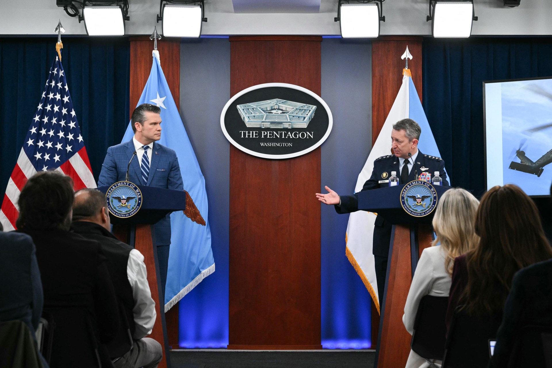 A military officer and civilian official speak at podiums during a press conference in The Pentagon, with the U.S. flag and two other flags displayed behind them.