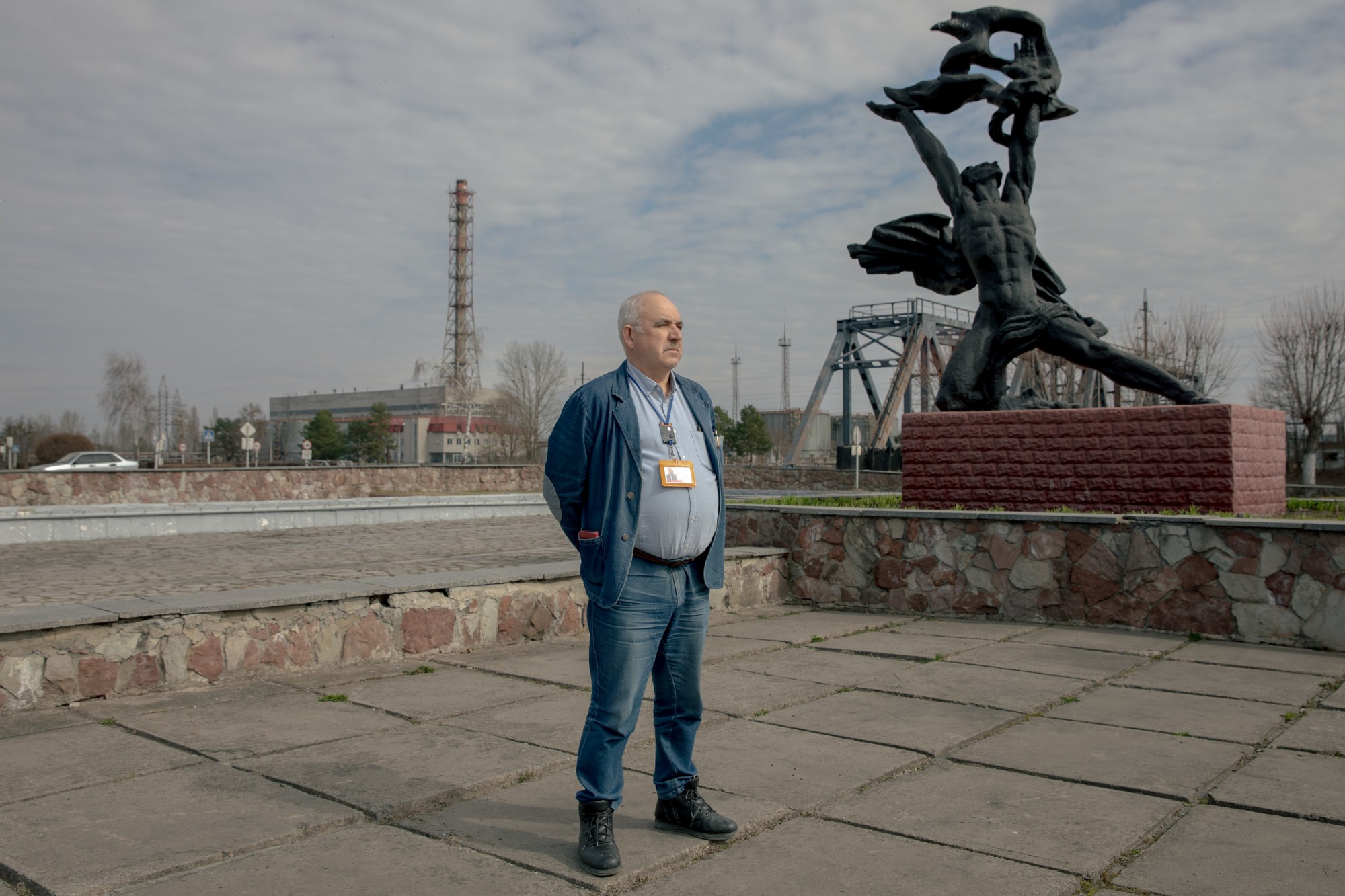 A man wearing a blue jacket and ID badge stands on a paved plaza next to a dramatic dark sculpture of a figure holding aloft a hammer and sickle, with industrial buildings and a river visible in the background.