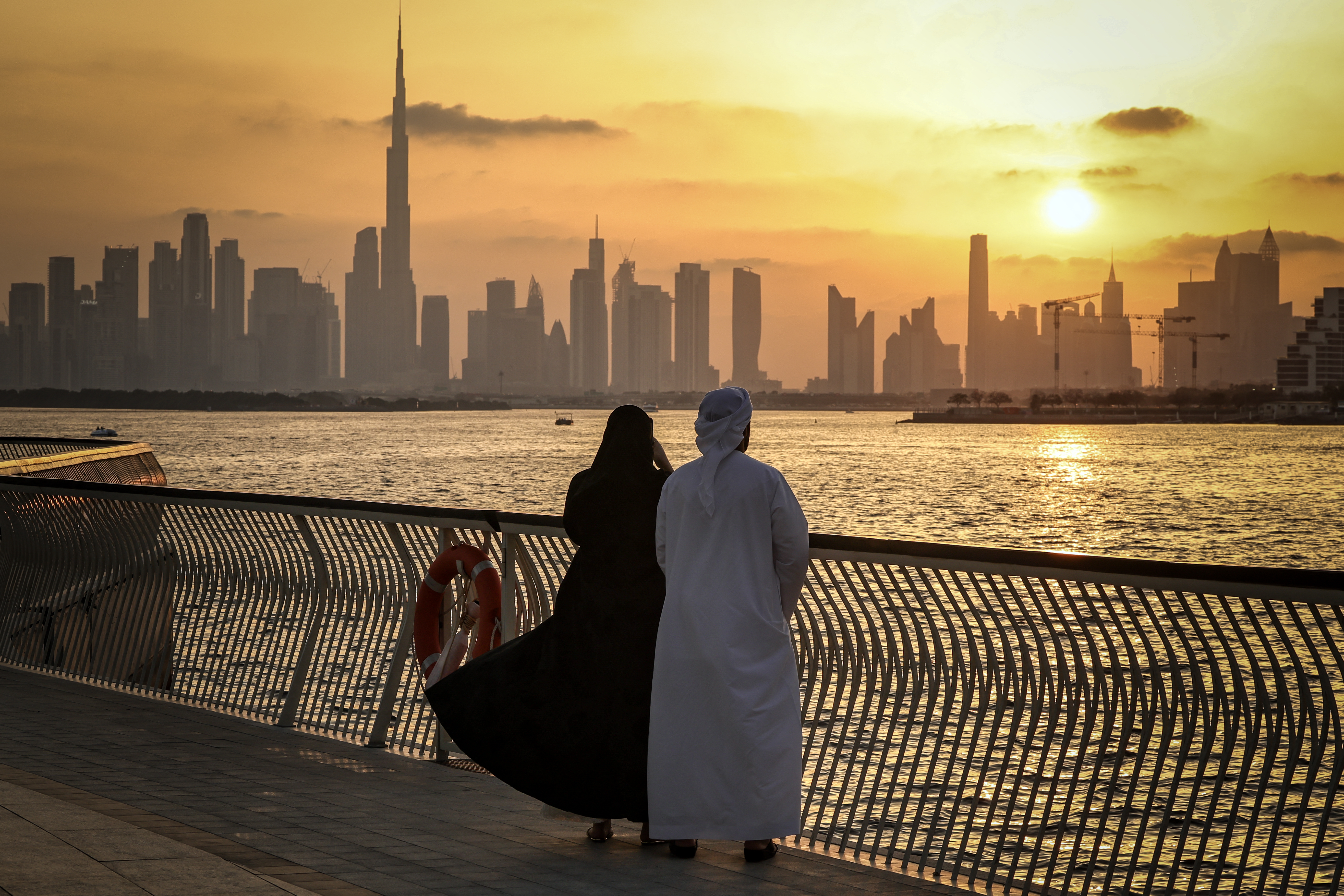 Two people in traditional Middle Eastern clothing stand at a waterfront railing, gazing at a city skyline during a golden sunset.