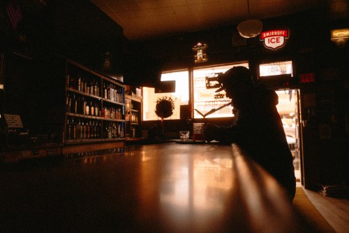 A silhouetted person stands at a bar counter in a dimly lit establishment with a Smirnoff Ice neon sign, bottle-lined shelves, and glowing storefront windows.