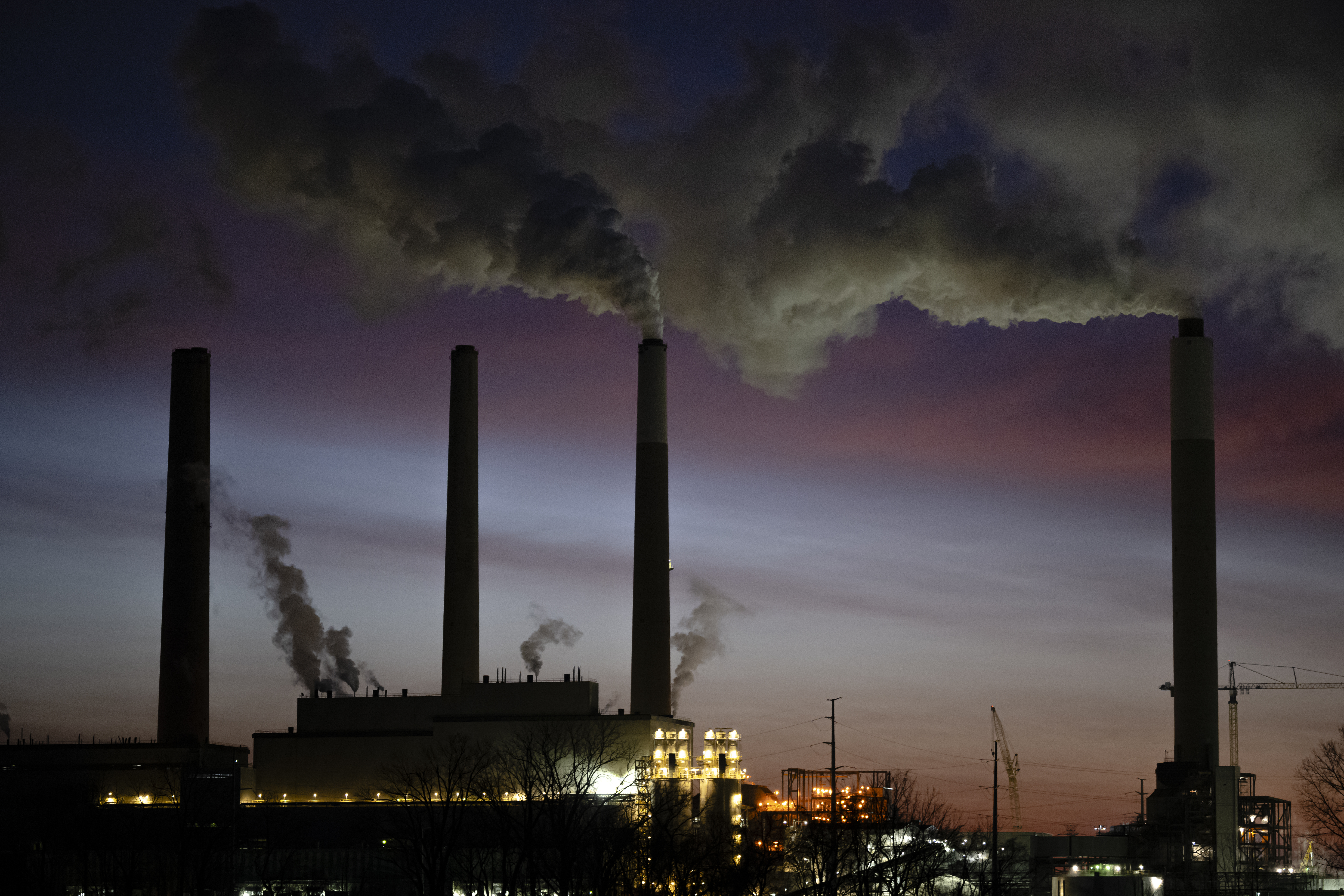Industrial power plant with four tall smokestacks silhouetted against a twilight sky, emitting dark smoke into clouds at dusk.