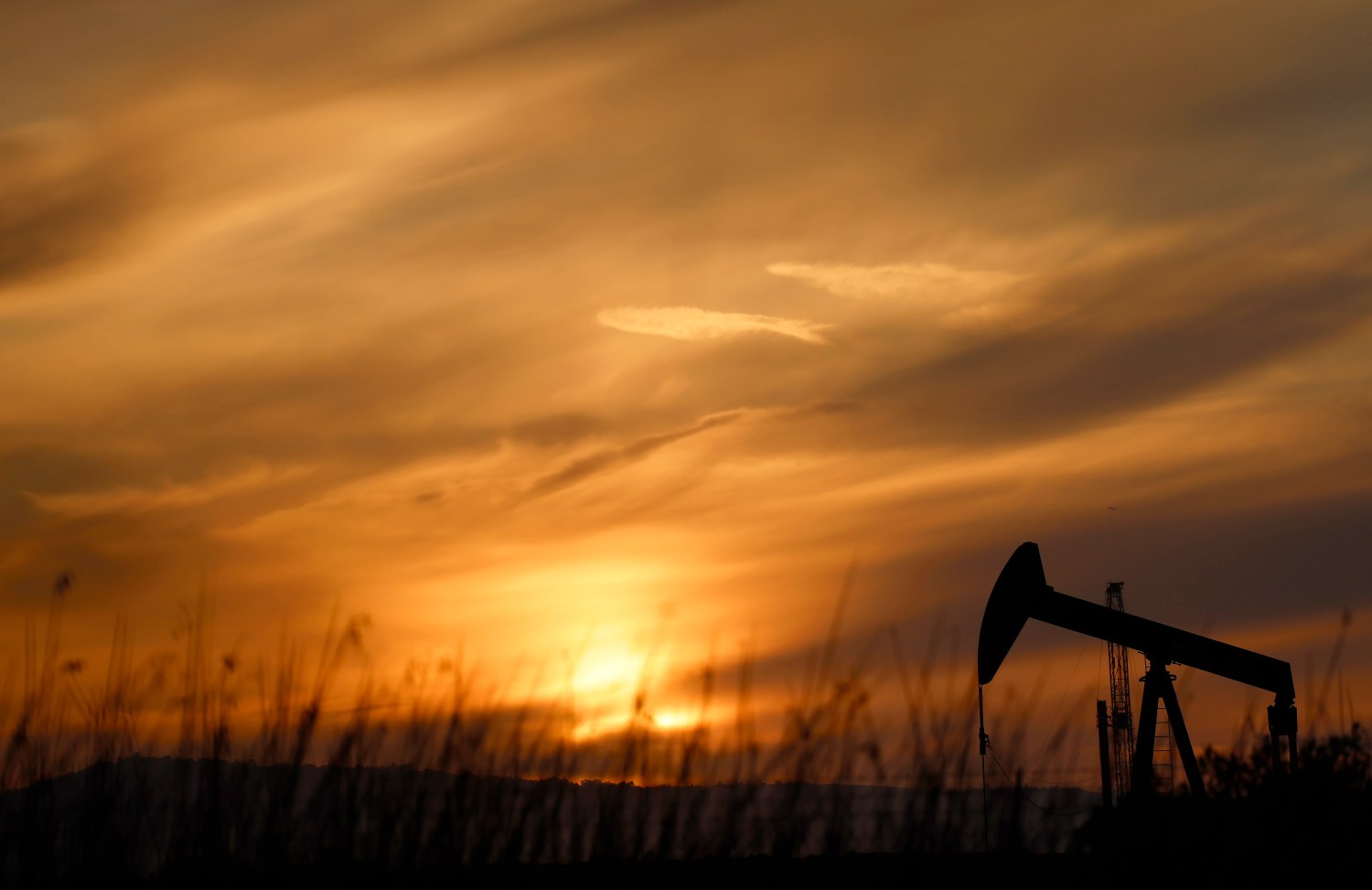 An oil pump jack silhouetted against a golden sunset in an arid landscape with sparse vegetation.