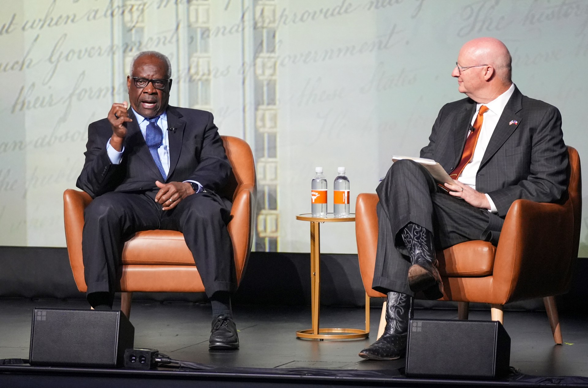 Two men in business suits sit in orange leather chairs on a stage during a formal discussion or interview, with water bottles and a small table between them, against a backdrop featuring historical architecture and text.