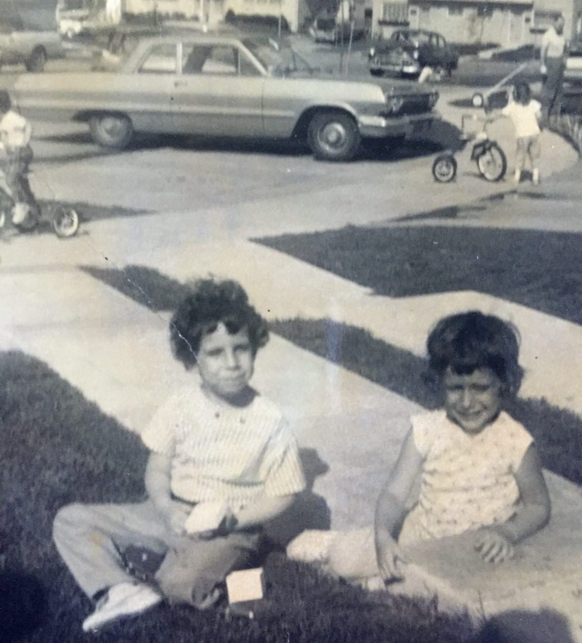 Two children sit on a driveway in a residential neighborhood while other children play with bicycles and cars in the background, circa 1950s.